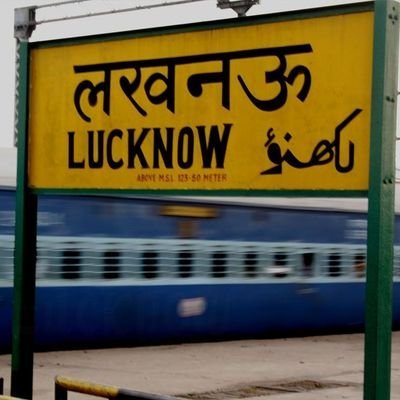 the name “LUCKNOW” written in English, Hindi, and Urdu scripts. The signboard is yellow with black text and is mounted on two green poles. Behind the signboard, a blue train carriage is visible, indicating that this image was taken at a railway station.