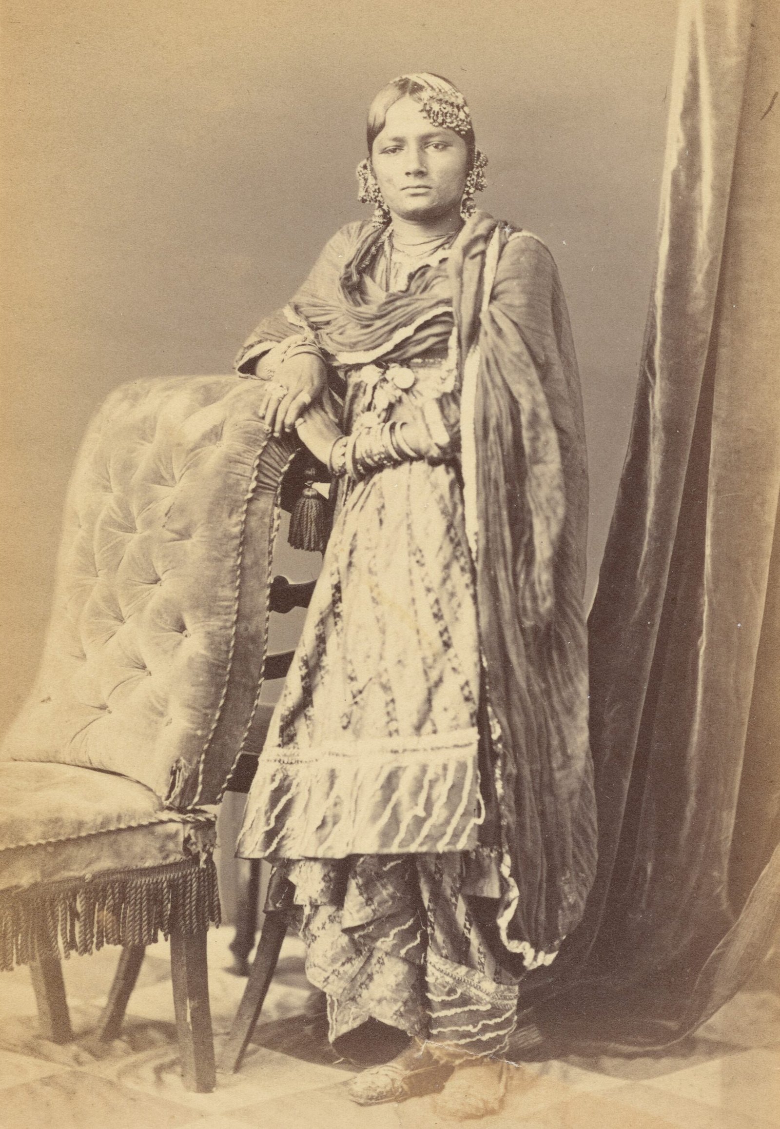 a vintage, sepia-toned photograph of a person standing next to an upholstered chair. The person’s face is obscured by a blank rectangle, so their identity and expressions are not visible. They are adorned in elaborate attire, including a detailed dress and shawl. The image has a sepia tone, indicating it is old or styled to appear vintage. The individual is wearing an elaborate dress with intricate designs and patterns. A shawl or draped fabric covers the shoulders and arms of the person. The chair next to which the person stands is plush, with tufted upholstery and fringed bottom edges. There’s a curtain in the background that appears heavy and rich in texture a lady wear gharara