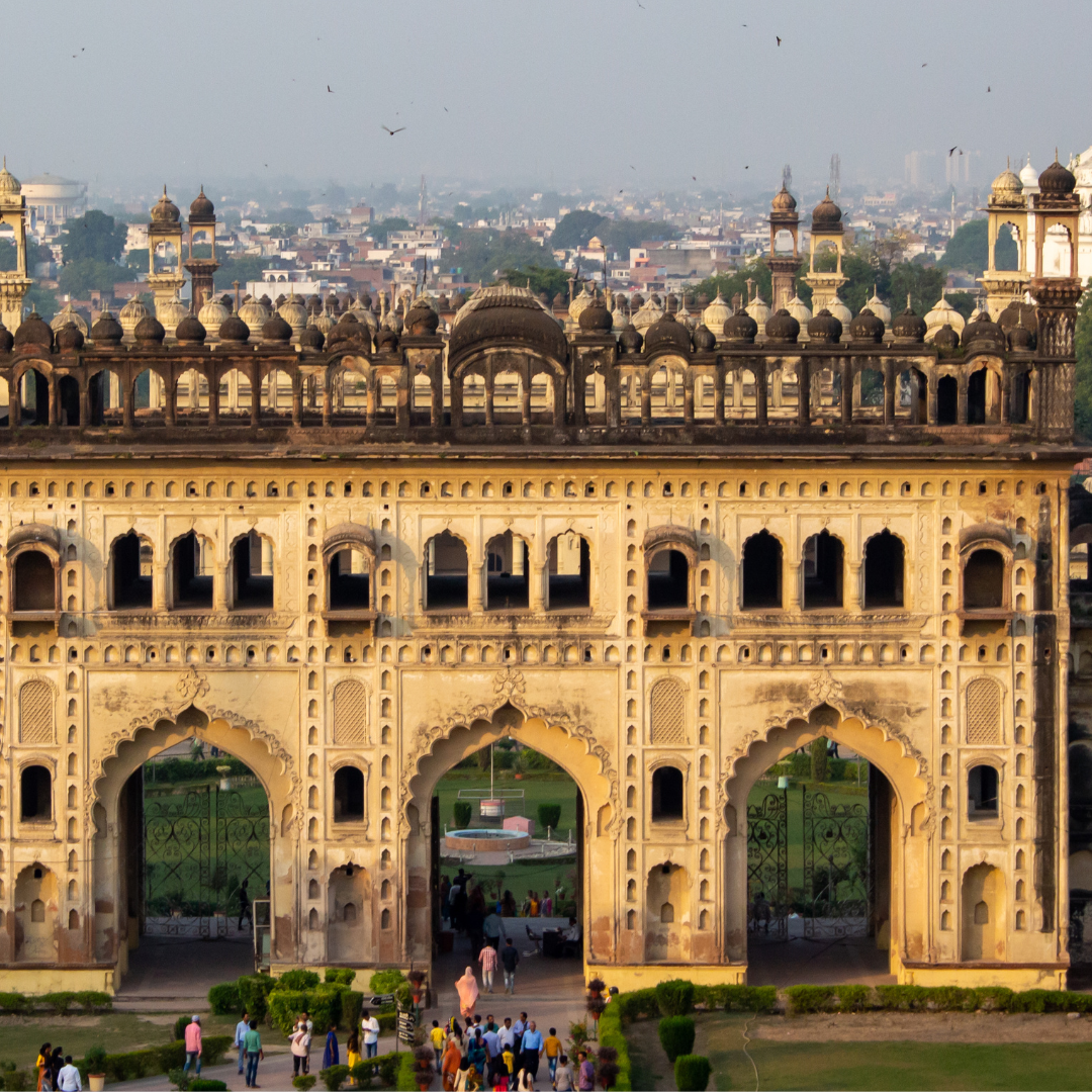 a grand, historic structure with intricate architectural designs. It features multiple arches and domes, adorned with detailed carvings and decorations. The building is made of a beige-colored stone or concrete material, giving it an ancient, majestic appearance. A group of people can be seen at the base of the structure, indicating its large scale. In the background, a cityscape under a clear sky is visible.