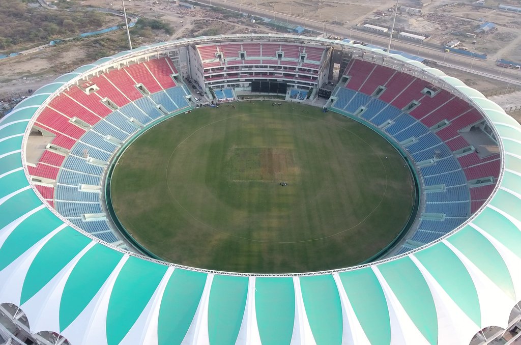 a large, oval-shaped stadium with a lush green field in the center. The seating area is divided into sections with different colors, including red and blue. The exterior of the stadium is adorned with white and turquoise stripes, giving it a modern aesthetic. Surrounding the stadium, there are open spaces and some construction or industrial areas visible. There’s no ongoing match or event as the field and seats are empty.