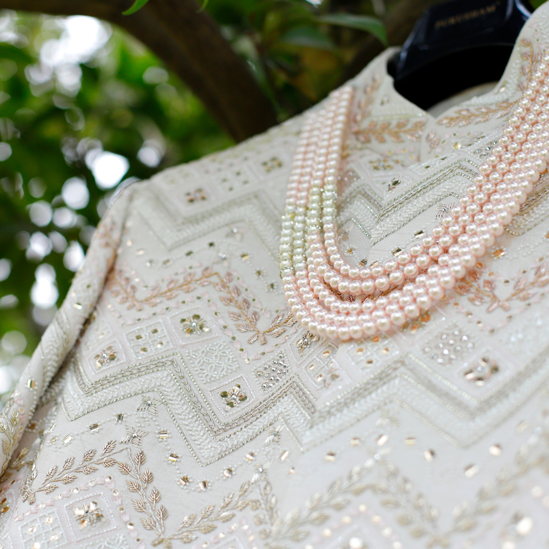 The central focus is on an intricately designed garment with detailed embroidery. Geometric and floral patterns adorn the fabric, enhanced by shiny embellishments. Strands of pearls in varying shades are draped elegantly across the garment. The fabric appears rich and luxurious, indicative of a formal or ceremonial attire. In the background, blurred foliage suggests an outdoor setting with natural lighting illuminating the details of the embroidery and pearls.