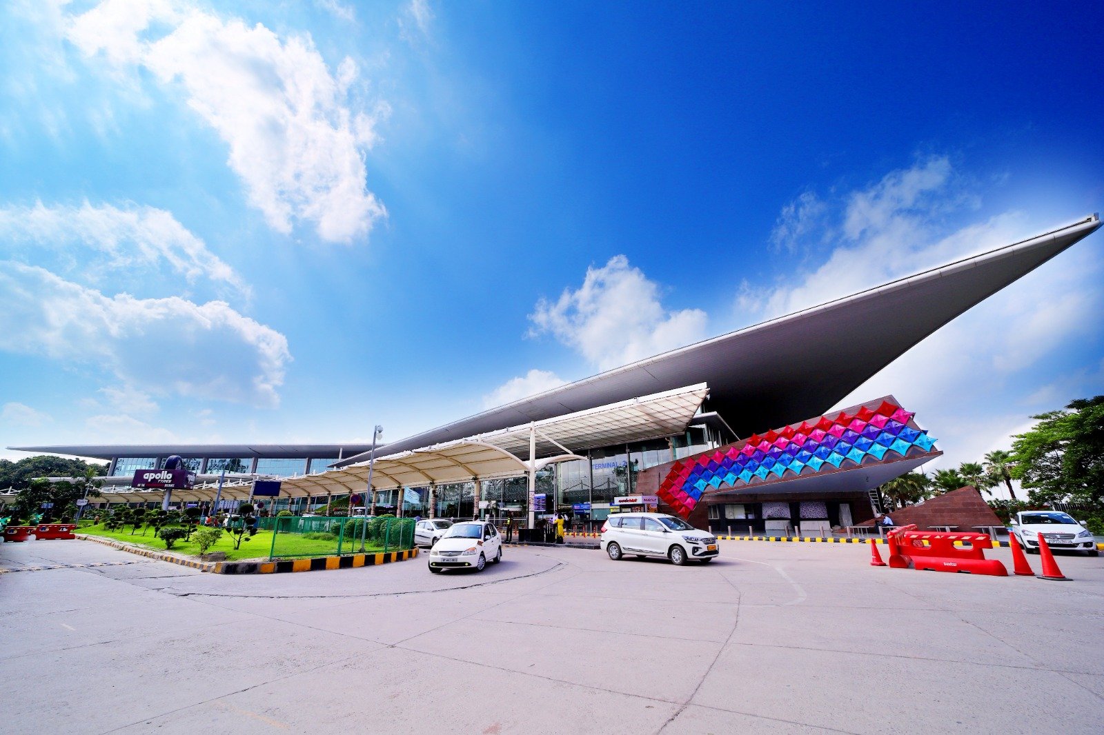 The image captures a modern architectural structure, likely an airport or a shopping mall, with a spacious parking area in the foreground under a bright blue sky with scattered clouds. The building has a sleek and modern design, characterized by its large overhanging roof and extensive use of glass. The parking area is spacious and well-maintained, with two white cars parked. There are colorful installations near the entrance of the building; one appears to be made of multiple colored cubes. A well-manicured lawn with small plants is visible beside the walkway leading to the entrance. The sky is bright and blue with scattered white clouds, indicating clear weather conditions.