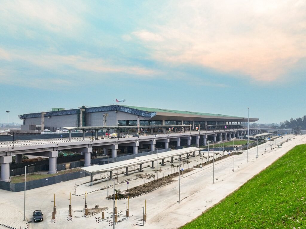 a large, modern airport terminal. The structure is elevated, with multiple lanes for vehicles underneath. The sky is partly cloudy.