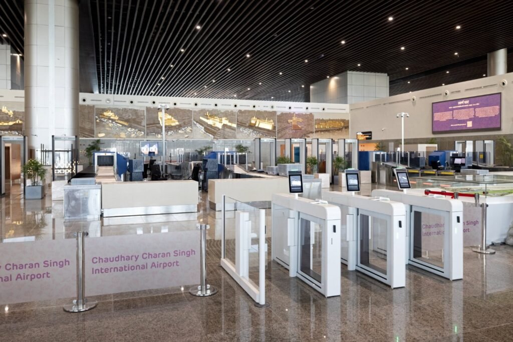 The image depicts an interior view of Chaudhary Charan Singh International Airport. The area is spacious and well-lit, featuring modern architecture and design.
The image shows the interior of Chaudhary Charan Singh International Airport.
There are multiple self-service kiosks with screens, placed in a row for passengers’ convenience.
Two large signs on the floor indicate the name of the airport.
In the background, there are counters with employees, possibly for customer service or check-in.
Above the counters, there is a large electronic display board showing flight information.
The ceiling is adorned with numerous small lights creating a starry effect.
The floor is made of polished stone or similar material, reflecting some of the overhead lights.