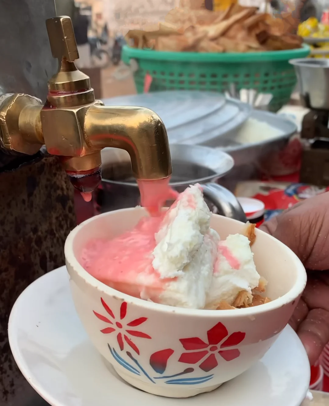 a moment where a pink syrup is being poured from a brass tap into a cup containing some form of dessert, possibly ice cream or whipped cream. The central focus is on the brass tap dispensing the pink syrup, which flows into a decorative cup adorned with red flower patterns. The cup sits on a matching saucer, held by a partially visible person’s hand. In the background, various items—including what appear to be fried snacks in green plastic baskets and other cooking utensils—suggest that this scene might be at a street food stall or an outdoor eating area.