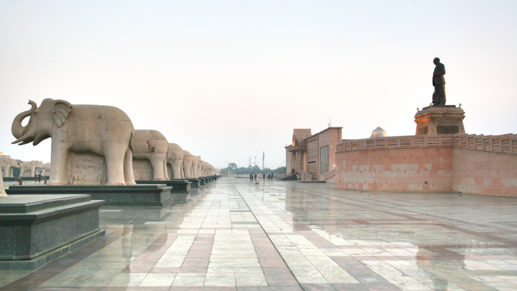 Eco Garden in Lucknow. Let’s explore the details:

Elephant Statues: On the left side, a row of large elephant statues stands tall. These majestic sculptures, with their trunks raised, create a sense of grandeur and reverence.
Promenade and Flooring: A wide promenade stretches ahead, adorned with intricately patterned flooring. The design adds an artistic touch to the pathway.
Central Statue: On the right, a raised platform hosts a statue of an individual. This figure stands upright, holding what appears to be a book or tablet. The pose suggests wisdom and contemplation.
Ambiance: The sky is overcast, casting a soft, diffused light. The absence of people in the area creates an empty and tranquil atmosphere—perfect for reflection and solitude.