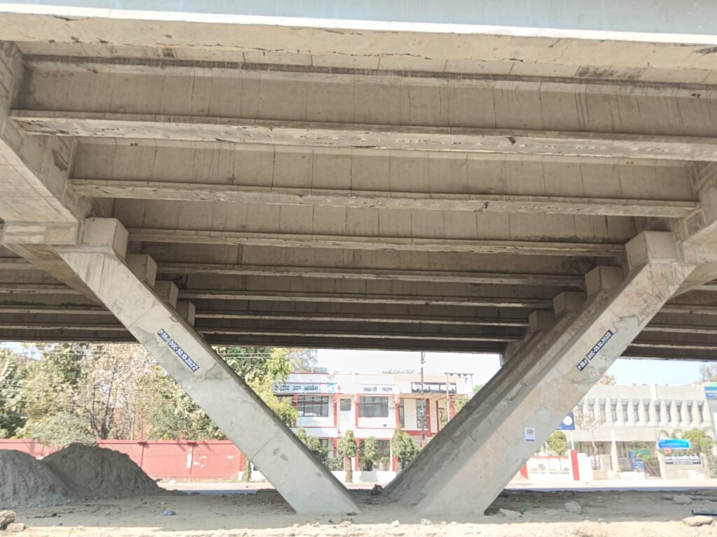 underside of a concrete bridge, revealing its structural elements and the space beneath it. The weathered grey concrete bridge features large, sturdy beams forming a V-shaped support. Below the bridge, an open area contains scattered dirt and debris. Urban structures are visible in the background under a clear sky. Blue rectangular signs are attached to two of the V-shaped supports, although their content remains unclear.