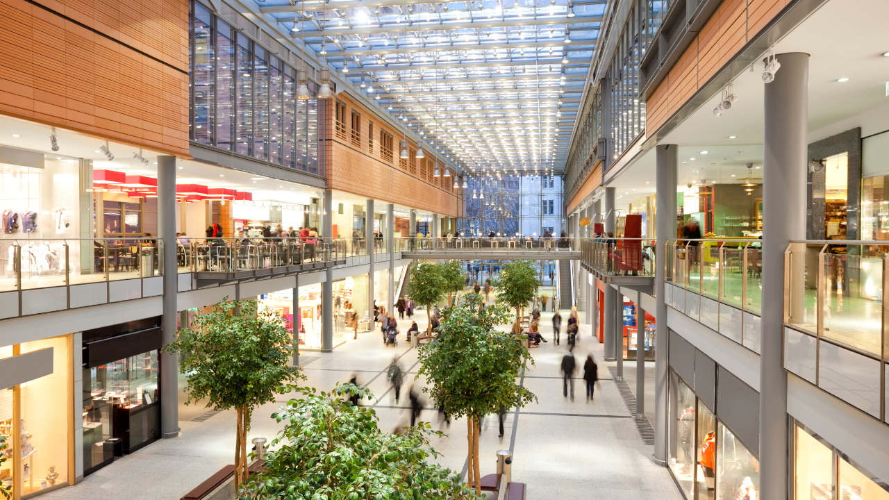 The mall features a wide central walkway flanked by multiple levels of stores on either side. Glass ceilings allow natural light to illuminate the interior, creating a bright and inviting atmosphere. Floors are connected by escalators located in the center of the view, providing easy access to different levels. Shoppers can be seen walking around, browsing the various stores and enjoying their shopping experience. Green potted plants add a touch of nature and freshness to the space. The mall’s architecture includes wooden panels and metal railings, contributing to a contemporary aesthetic.