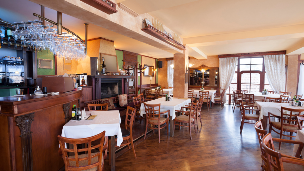 The room is spacious with multiple wooden tables, each set with white tablecloths, napkins, and cutlery.
Wooden chairs are neatly arranged around the tables.
To the left, there is a wooden bar counter with various bottles on shelves above it and glassware hanging from a rack.
A coffee machine is visible on the bar counter.
There’s a fireplace near the bar area, adding to the warm ambiance of the room.
The walls are painted in warm tones and decorated with small framed pictures.
Large windows allow natural light to fill the room, enhancing its welcoming atmosphere.