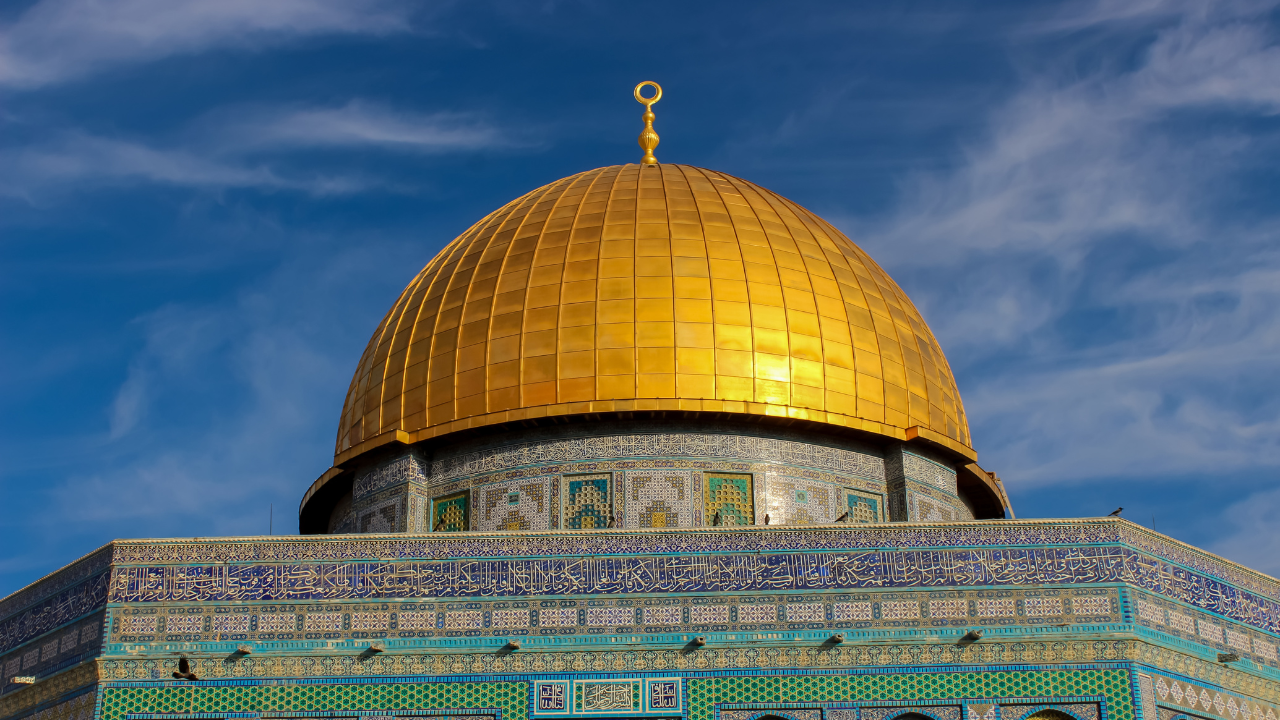 a majestic golden dome atop a significant religious or historical building. The dome’s reflective surface gleams under the sunlight, while intricate geometric patterns adorn its base. Below the dome, vibrant tile work features Arabic calligraphy and ornamental designs in shades of blue, turquoise, white, and gold. The clear blue sky serves as a serene backdrop, with scattered white clouds adding to the scene.