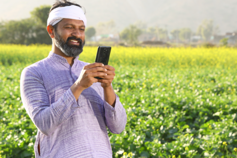 a person standing amidst a field of vibrant yellow flowering plants. They hold a smartphone, their face obscured by a blurred rectangular area for privacy. The rural setting, with greenery and distant hills, suggests a connection between technology and agriculture—a theme resonant with Gramik’s mission to empower Indian farmers.