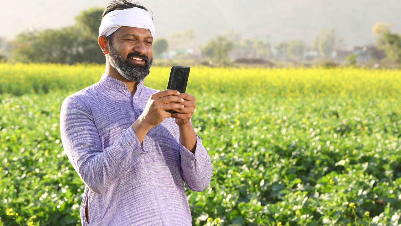 a person standing amidst a field of vibrant yellow flowering plants. They hold a smartphone, their face obscured by a blurred rectangular area for privacy. The rural setting, with greenery and distant hills, suggests a connection between technology and agriculture—a theme resonant with Gramik’s mission to empower Indian farmers.