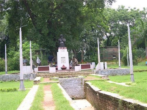 Central focus on a statue placed on a pedestal, which is the main subject of the image.
The statue is surrounded by multiple flagpoles without flags.
There are benches on either side of the paved pathway leading to the statue.
The area is lush with green grass and trees in the background, suggesting a park or memorial space.
The sky appears overcast, giving a calm ambiance to the setting.
