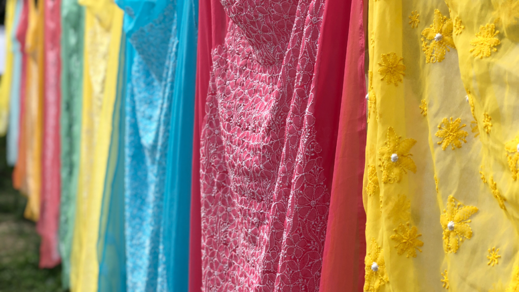 a vibrant collection of colorful fabrics hanging side by side. Each fabric piece boasts unique textures and patterns, ranging from delicate lace detailing to floral motifs. The natural light illuminates these fabrics, enhancing their vivid colors. Although the setting isn’t explicitly clear, the blurred background suggests an outdoor environment.