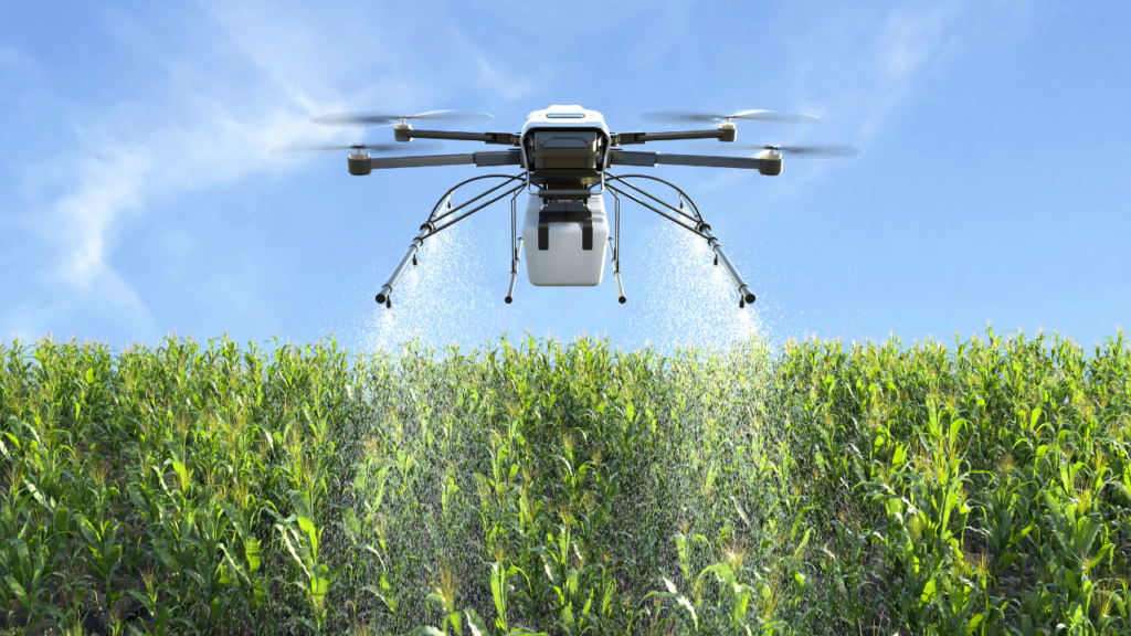 The image depicts a white drone with four rotors, hovering above a green field of crops under a clear blue sky.
The drone is equipped with two spraying nozzles, releasing a mist of liquid onto the plants below.
The lush green field of tall crops stretches out beneath the drone, indicating an agricultural setting.
The sky is clear and blue, with few clouds, suggesting good weather conditions for outdoor activities or work.
There’s an implication of agricultural technology being used for crop maintenance or protection.